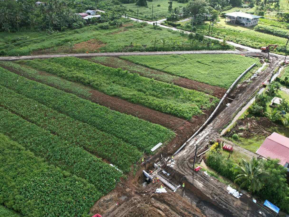 Overhead perspective of a farm landscape, showcasing a road and multiple houses surrounded by fields and crop. A pipeline is in progress of being laid in a trench.